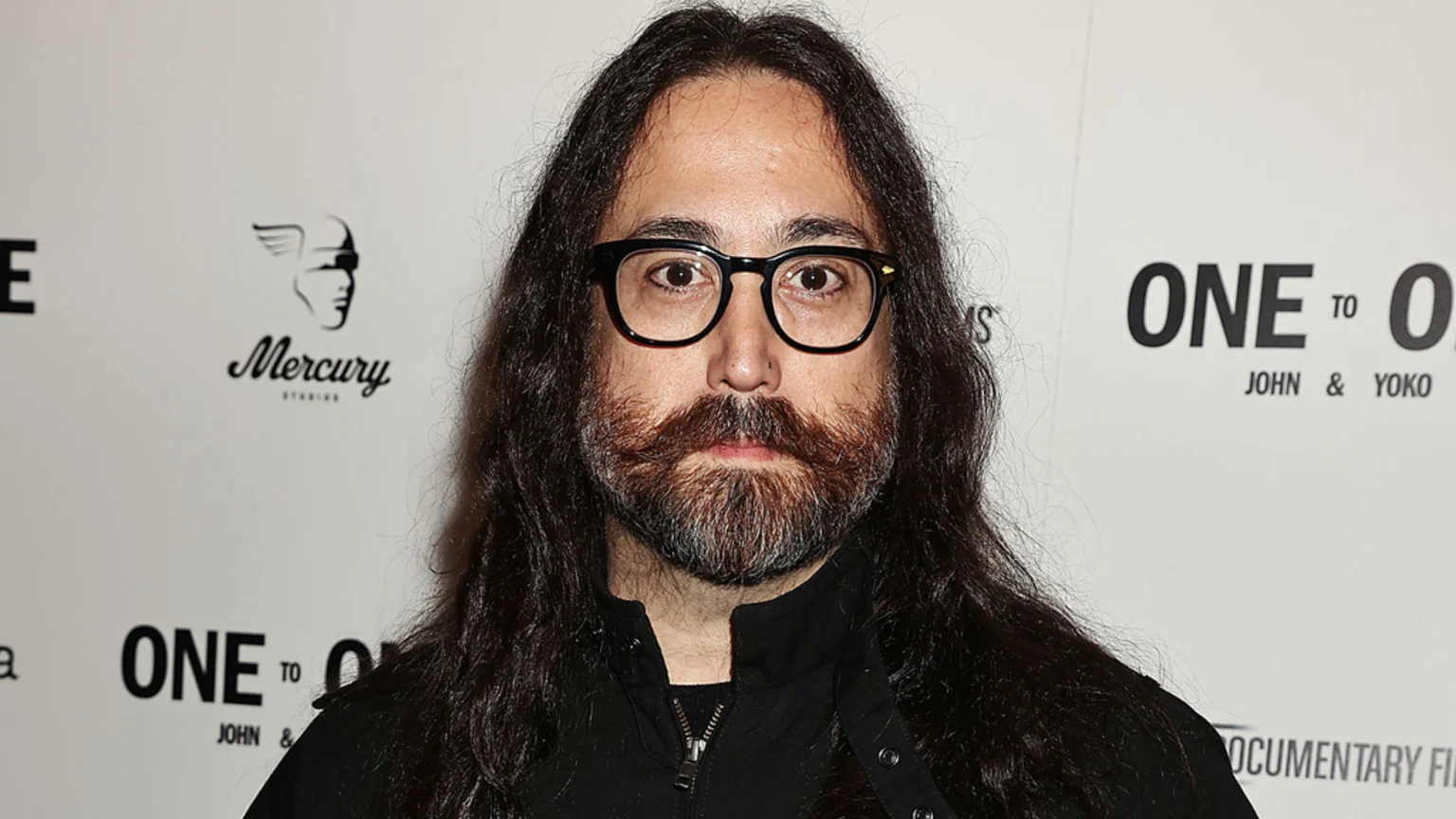 A man with long dark hair, a beard, mustache, and glasses looks forward, against a white background with "Mercury Studios" and "One to One John & Yoko" logos.