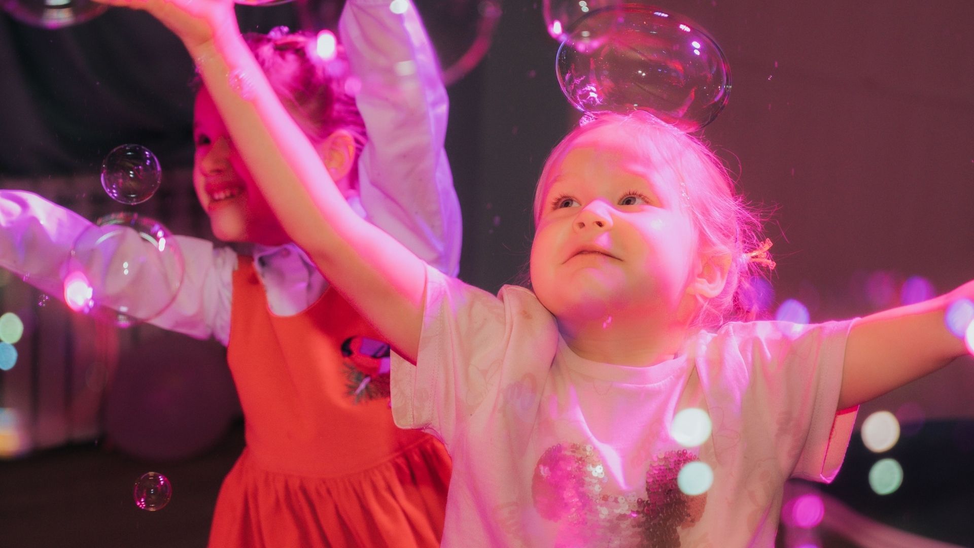 Two young children, bathed in pink and purple light, joyfully reach for numerous floating soap bubbles, with one child in the foreground gazing upwards, arms outstretched.