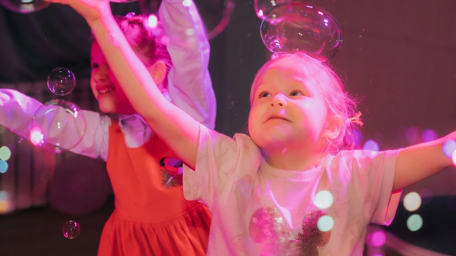Two young children, bathed in pink and purple light, joyfully reach for numerous floating soap bubbles, with one child in the foreground gazing upwards, arms outstretched.