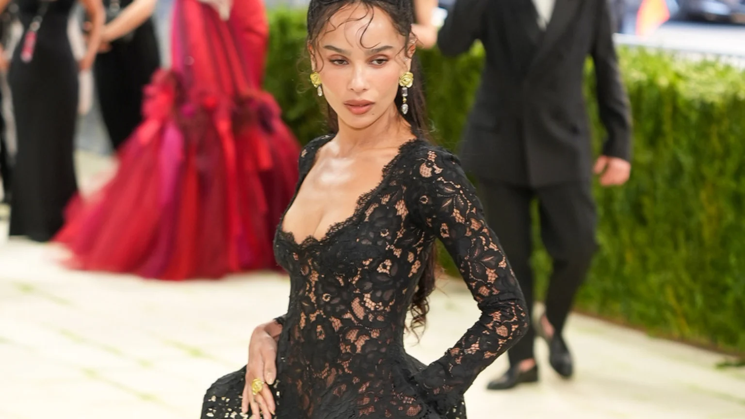 A woman in a black lace dress with a low neckline and long sleeves looks down, wearing ornate earrings and a large yellow ring, with blurry formal figures in the background.