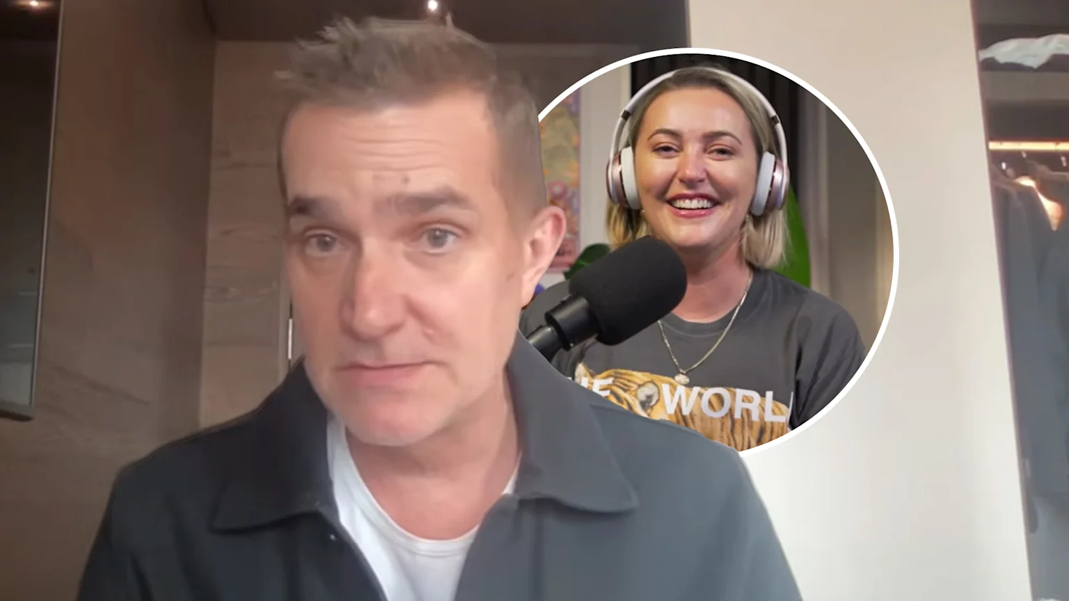 John Aiken, wearing a dark collared shirt, looks forward, as a smiling blonde woman in headphones and a tiger-print shirt speaks into a microphone in a circular inset.