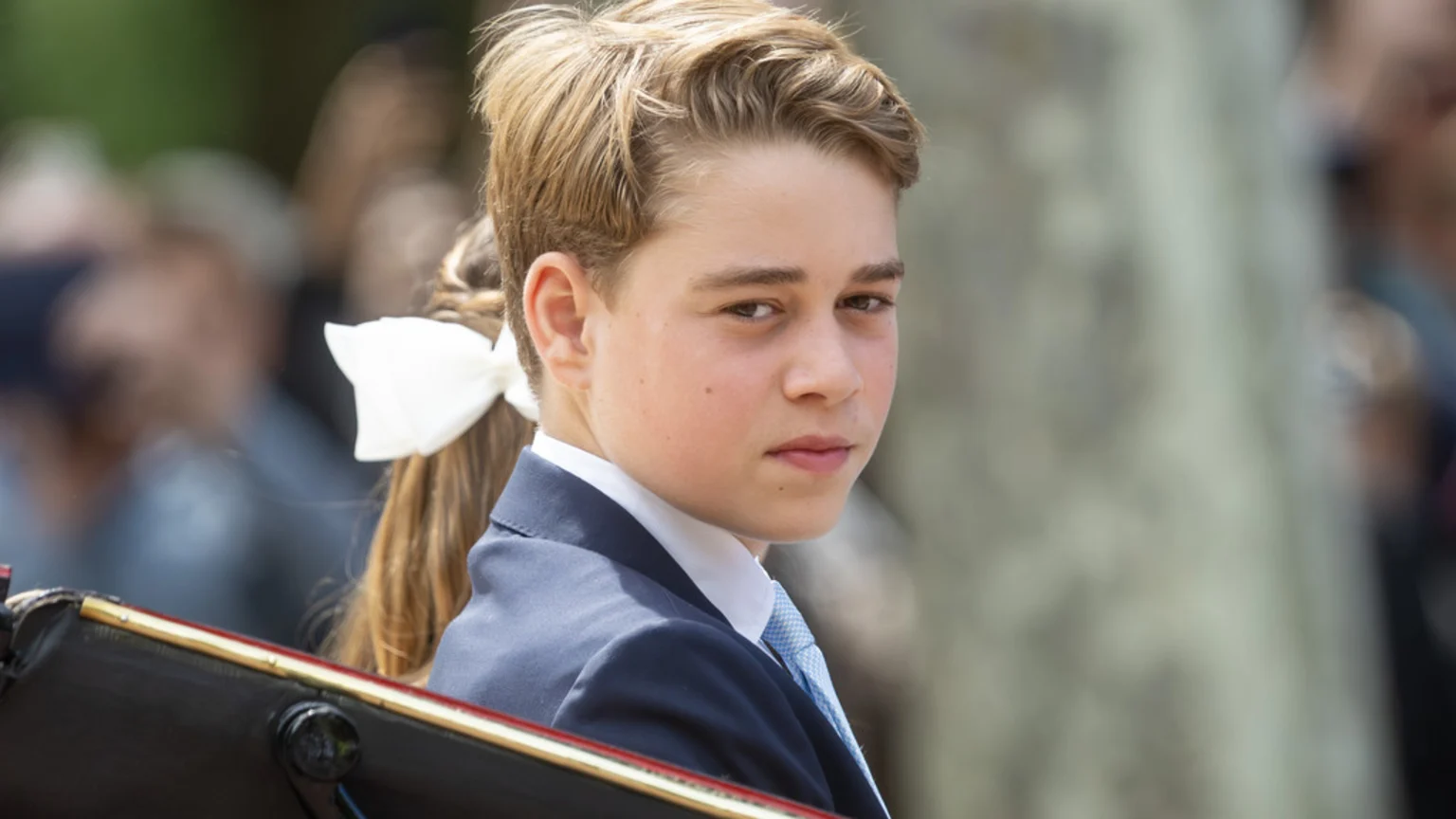 A young boy with light brown hair, wearing a navy suit and light blue tie, looks over his right shoulder with a serious expression, with a white bow visible in hair behind him.