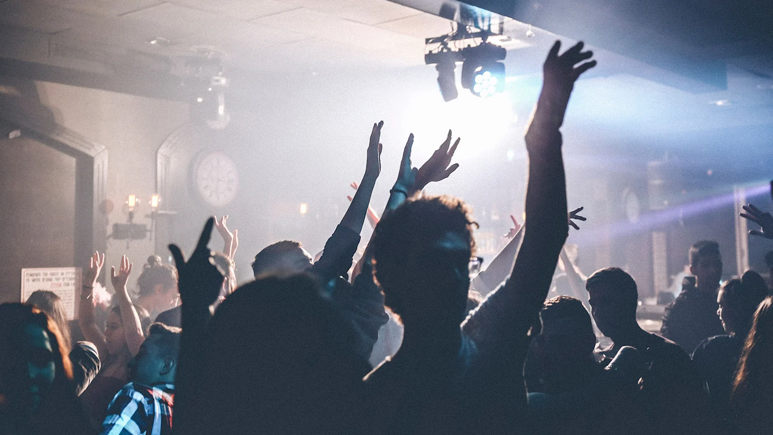 A dimly lit indoor scene shows a crowd of people, many with their arms raised, illuminated by bright spotlights cutting through a hazy atmosphere.