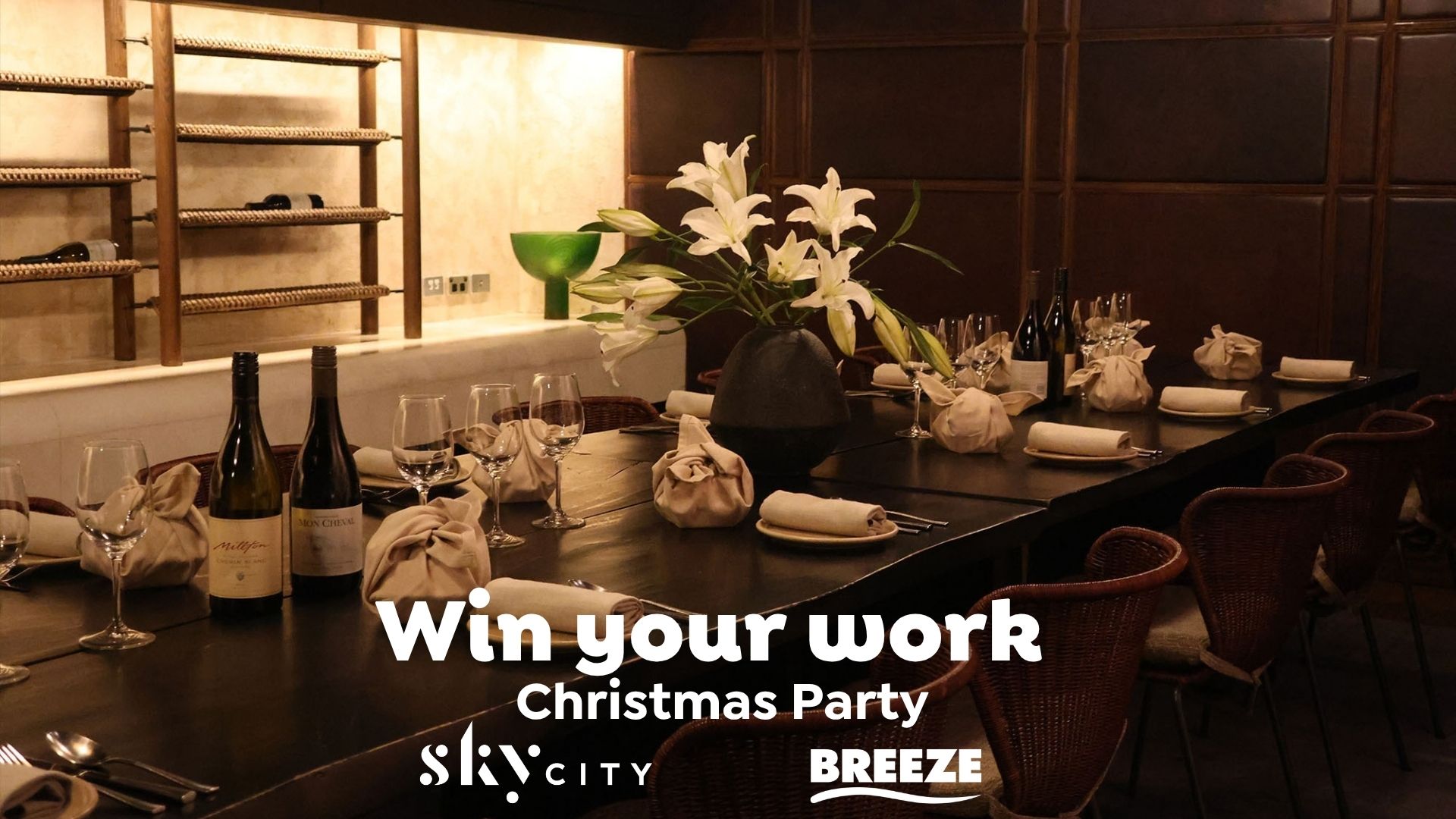 A long dining table is set with wine bottles, glasses, plates, and wrapped items, featuring a white lily centerpiece and wine racks in the background, with text "Win your work Christmas Party Sky CITY BREEZE".