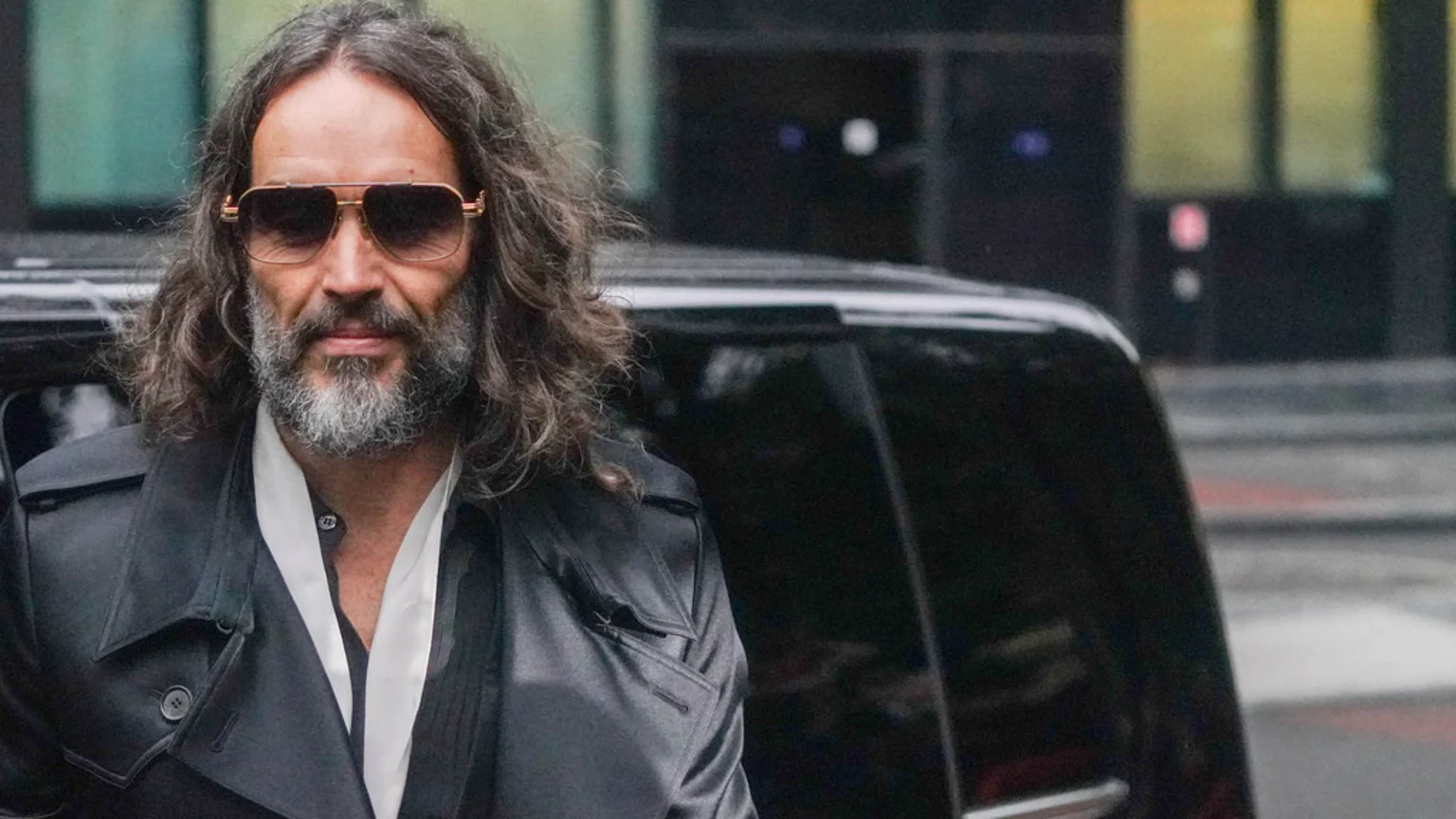 Russell Brand with long, wavy hair and a grey beard, wearing gold-framed sunglasses and a black trench coat over a white shirt, looks forward with a dark vehicle behind him.