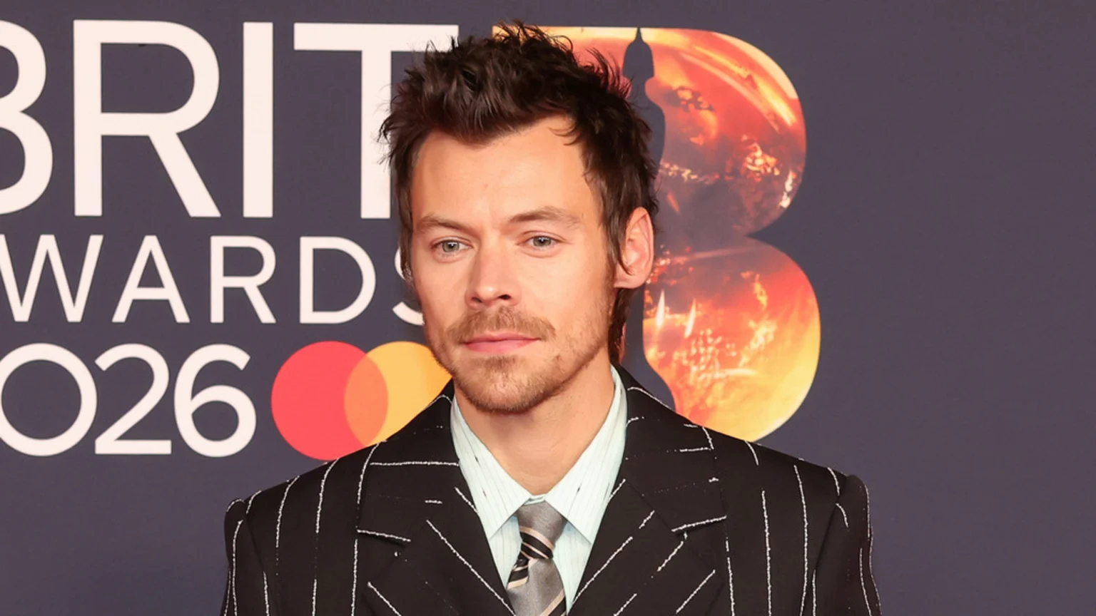 Harry Styles, a man with brown hair and stubble, stands in a pinstripe suit, light blue shirt, and striped tie, against a dark banner displaying "BRIT AWARDS 2026" and orange circular graphics.