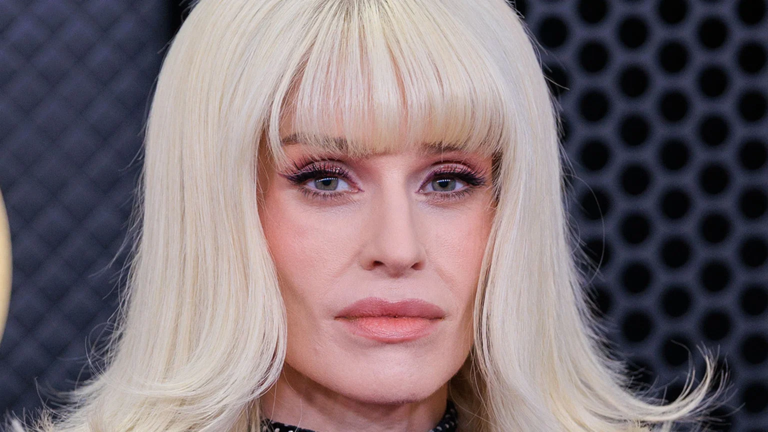 A woman with platinum blonde hair and bangs, wearing dramatic pink winged eye makeup, looks intently at the camera against a dark, textured background.