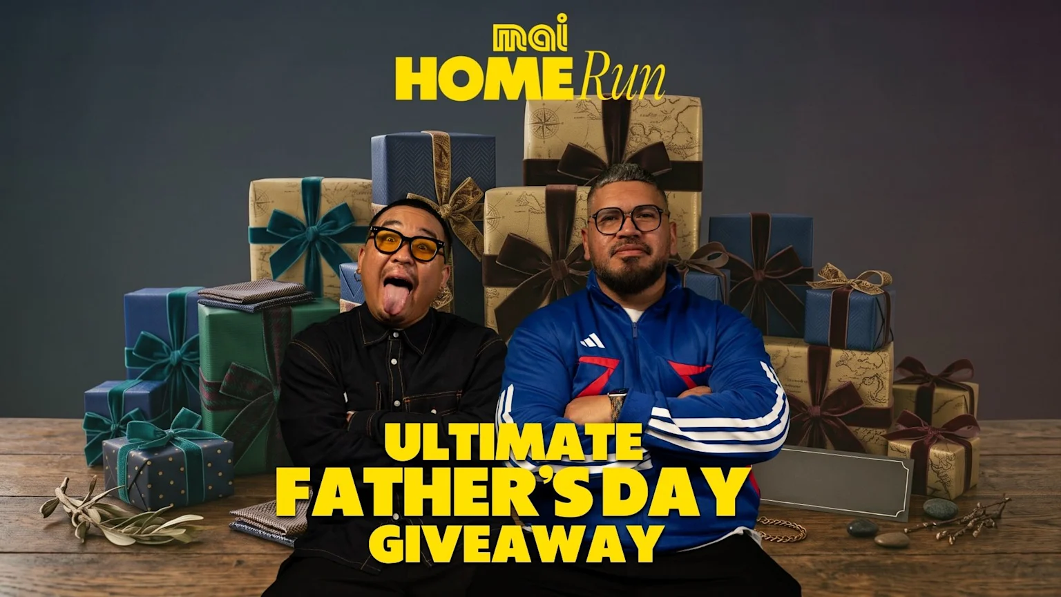 Two men, one making a funny face with his tongue out, and the other with a beard and crossed arms, sit in front of stacked gift boxes on a wooden table, with "mai HOME Run" and "ULTIMATE FATHER'S DAY GIVEAWAY" text.