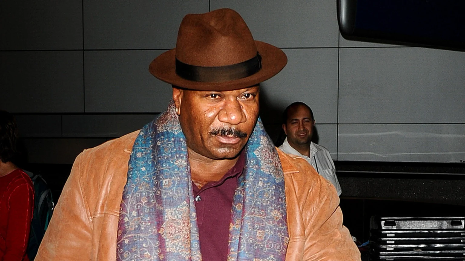 A man with dark skin, a brown fedora, and a patterned scarf, wearing a tan leather jacket and a burgundy shirt, looks slightly right while another man smiles in the background.