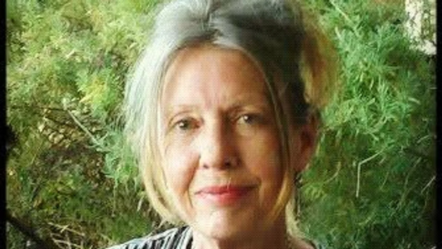 A portrait of an older woman with light gray and blonde hair, smiling subtly against a blurred green leafy background.