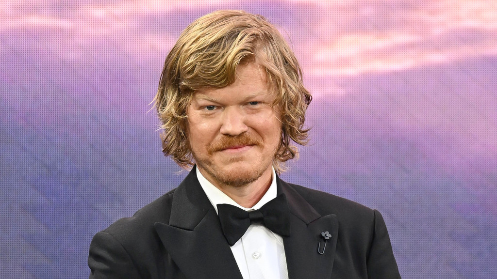 Jesse Plemons, with wavy blonde hair and a mustache, wears a black tuxedo with a bow tie and a floral lapel pin, smiling slightly against a blurred purple and pink background.