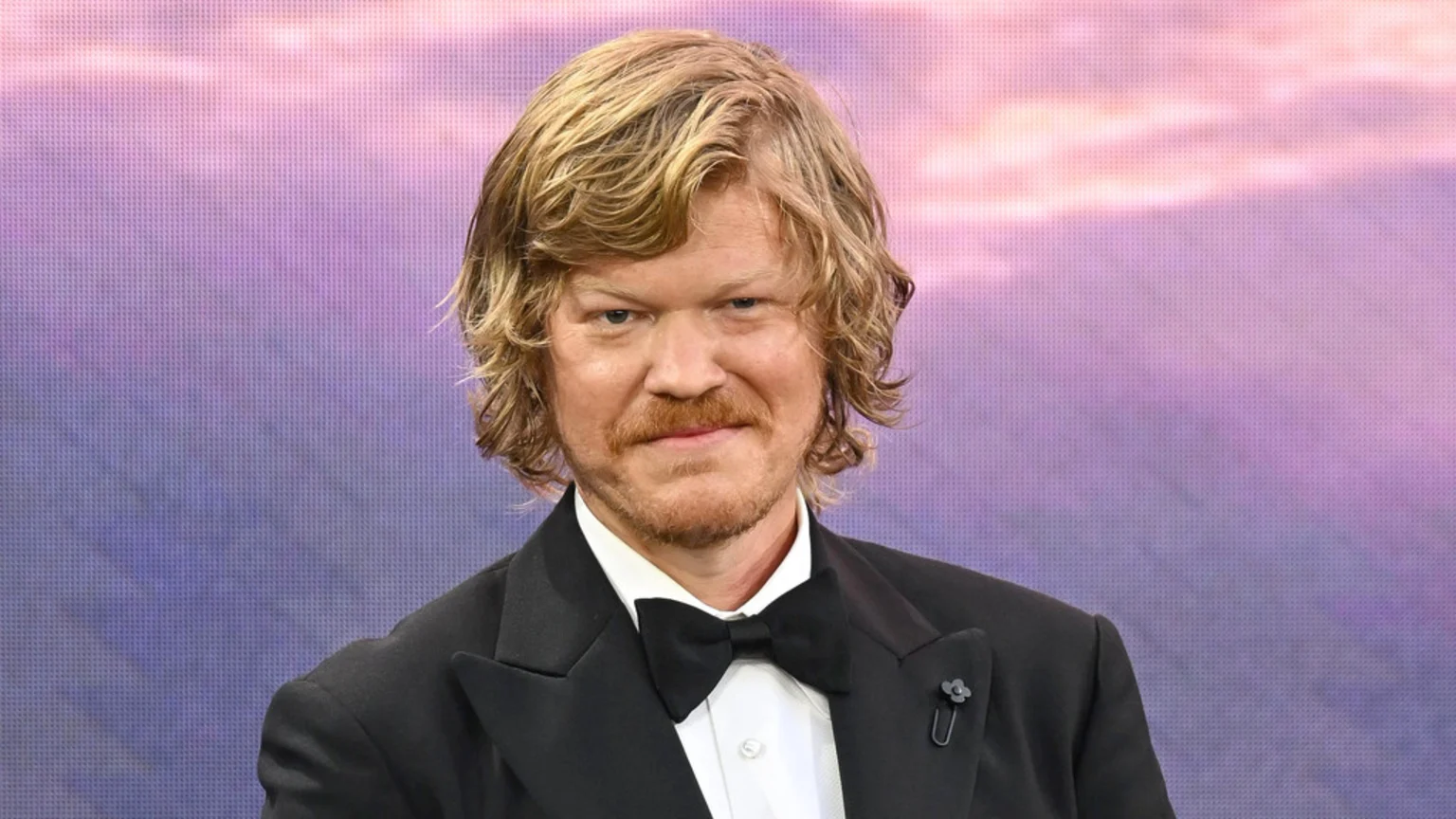 Jesse Plemons, with wavy blonde hair and a mustache, wears a black tuxedo with a bow tie and a floral lapel pin, smiling slightly against a blurred purple and pink background.