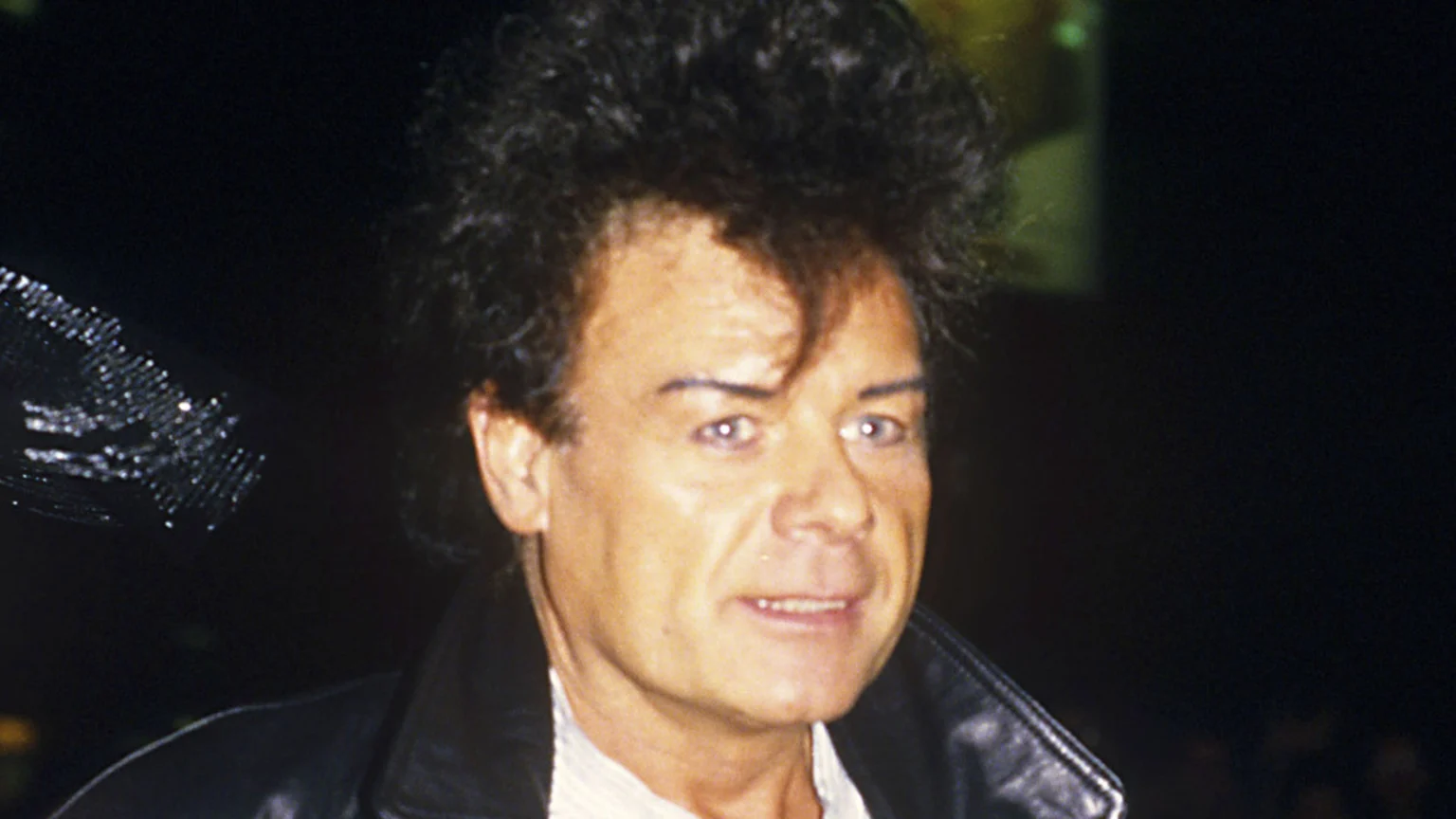 A man with prominent, dark, voluminous curly hair, thin eyebrows, and light eyes looks towards the viewer, smiling slightly, wearing a dark leather jacket over a light shirt against a dark background.
