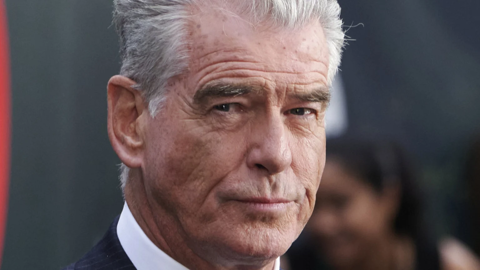 A close-up headshot of Pierce Brosnan, an older man with grey hair, looking directly at the camera with a serious expression, wearing a white shirt and dark pinstriped suit jacket.