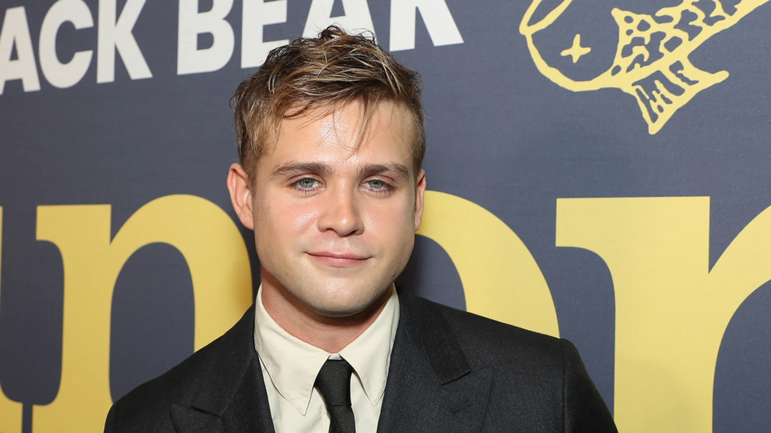 Actor Leo Woodall, a young man with light brown hair and blue eyes, wearing a dark suit, light shirt, and dark tie, smiles subtly against a dark blue background with yellow text and a fish graphic.