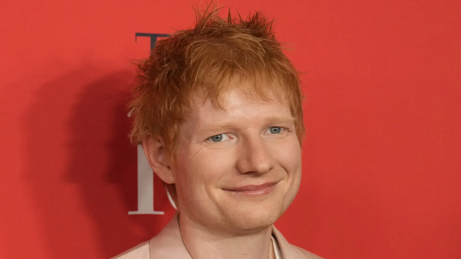 Ed Sheeran, a man with red hair and blue eyes, smiles while wearing a light suit against a red background with a partial 'TIME' logo.