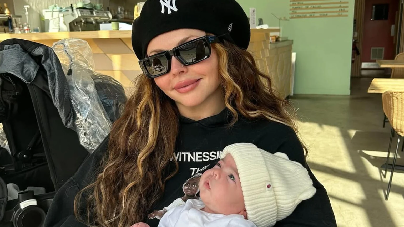 Jesy Nelson, wearing a black beret and sunglasses, smiles while cradling a baby in a white hat who looks up at her, with a stroller and a cafe interior visible in the sunlit background.