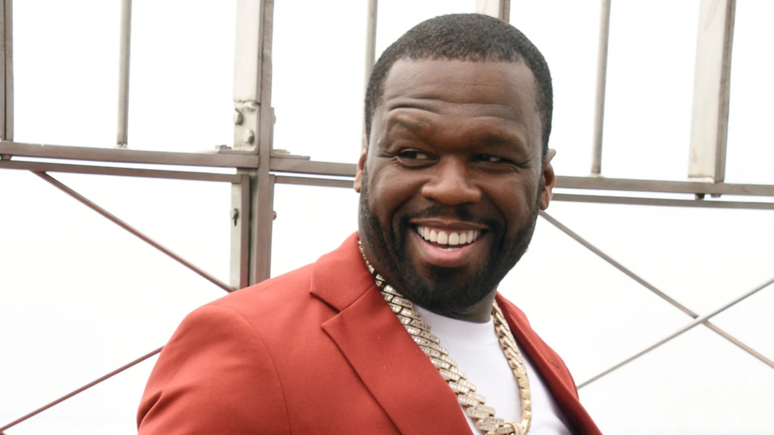 50 Cent, a Black man, smiles widely wearing a rust-colored jacket and a thick diamond chain, with metal structures in the background.