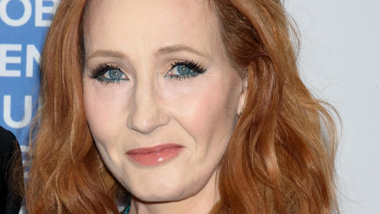Close-up of J. K. Rowling, a woman with red hair and blue eyes looking forward, wearing subtle makeup, with a blurred blue background.