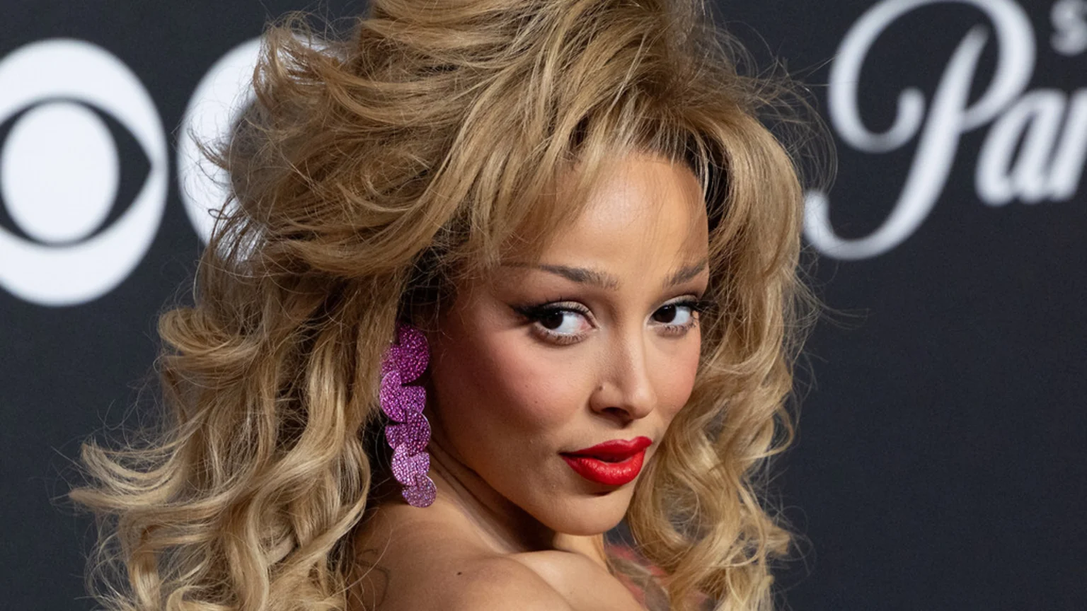 A woman with blonde, voluminous hair and bangs looks over her shoulder, wearing red lipstick, winged eyeliner, and pink disc earrings, against a dark background with blurry white logos.
