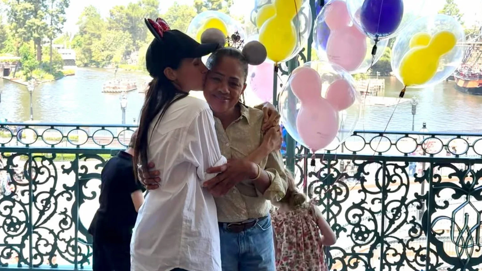 A woman wearing Minnie Mouse ears kisses a woman wearing Mickey Mouse ears on the cheek while they embrace, surrounded by Mickey-shaped balloons, a railing, and a park with water and boats in the background.