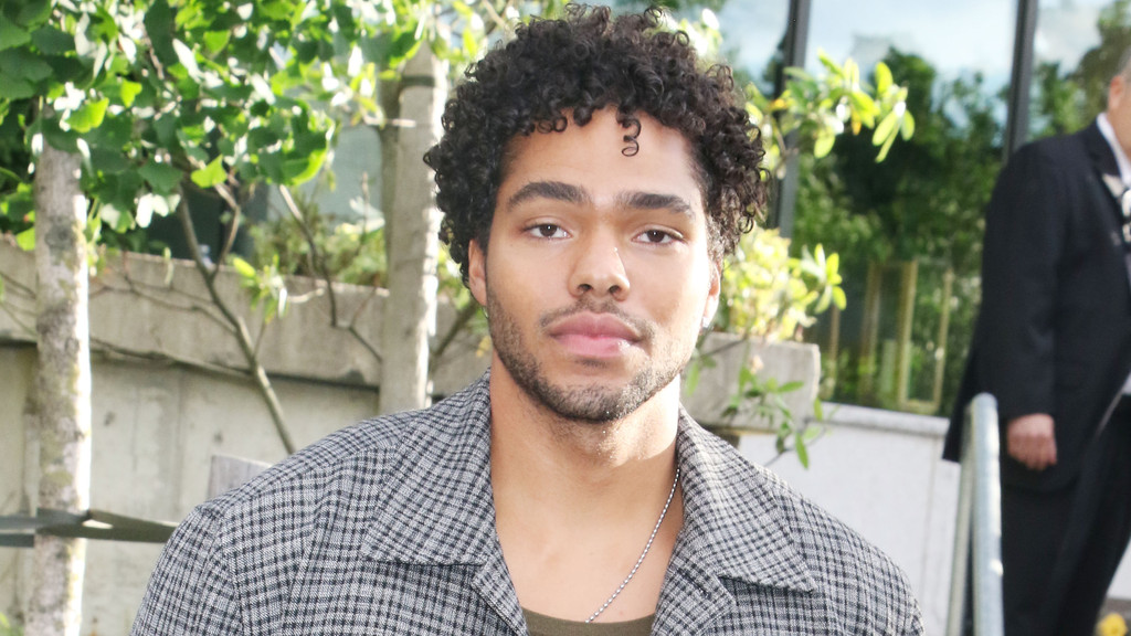 Close-up of actor Nicholas Duvernay, a young man with curly hair and a beard, wearing a plaid jacket, looking at the camera outdoors.