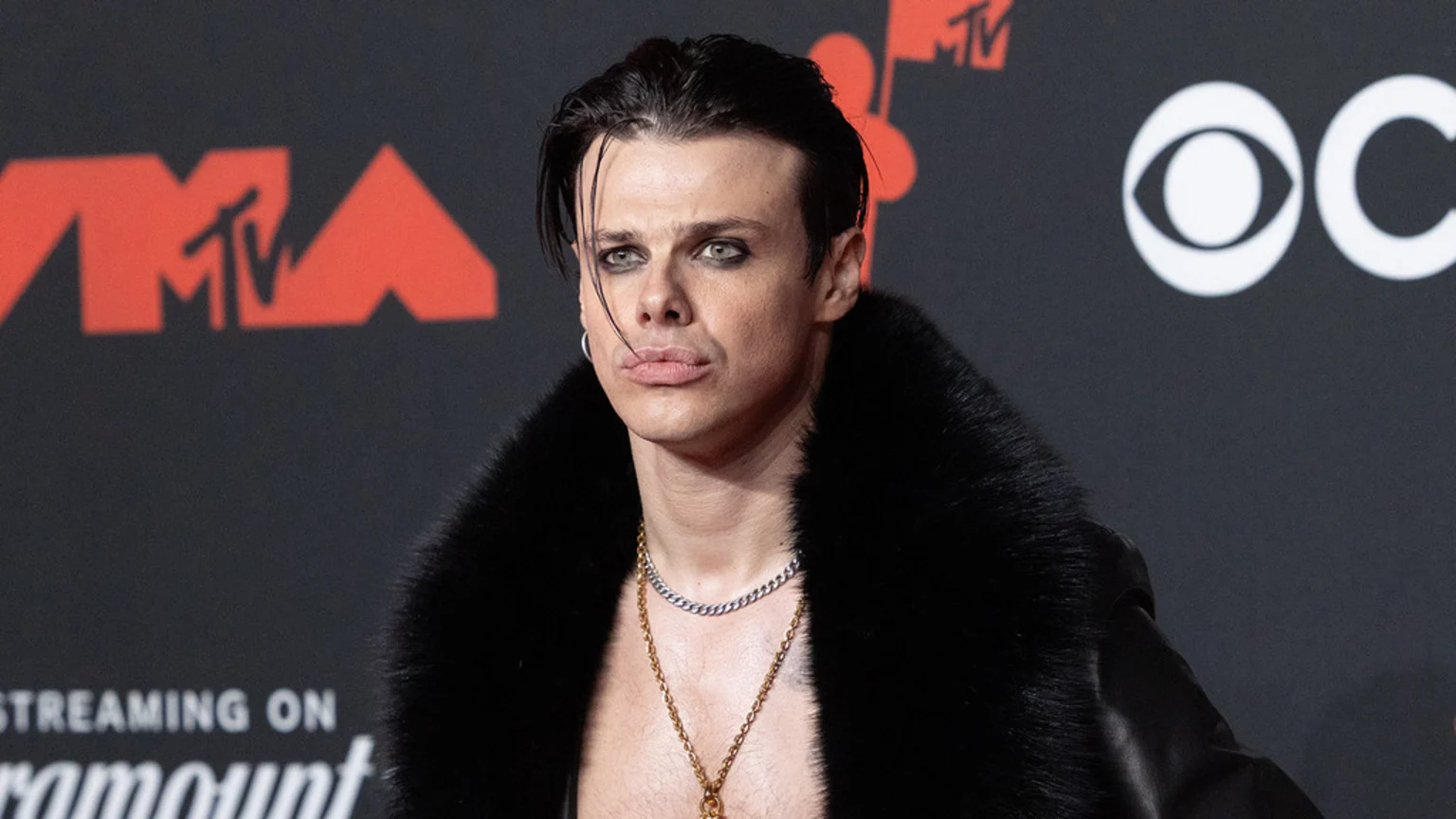 Yungblud, wearing dark eye makeup and an open black fur coat over his bare chest, poses against a red carpet backdrop featuring MTV and CBS logos.