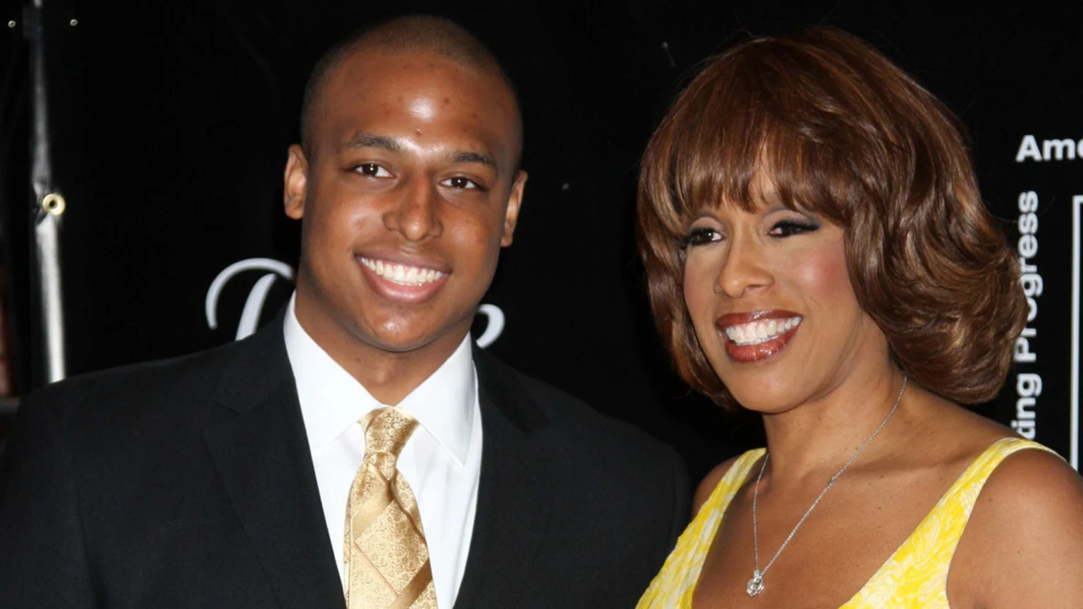 A smiling man in a black suit and a woman in a yellow dress with reddish-brown hair stand side-by-side against a dark background.