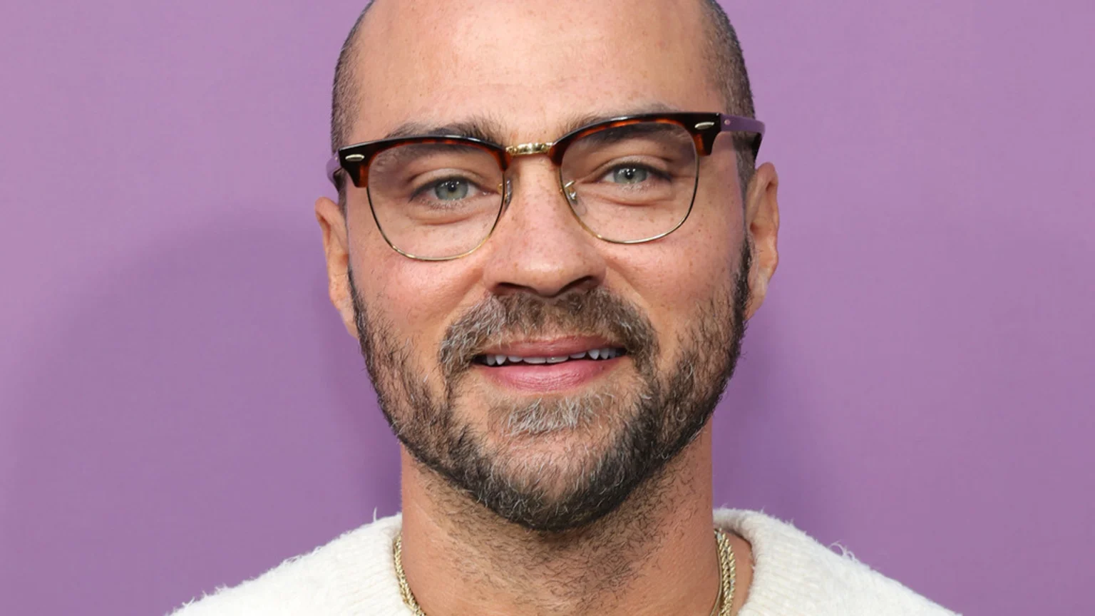 A man with a beard and glasses smiles faintly, wearing a light-colored fuzzy sweater and a gold chain against a purple background.