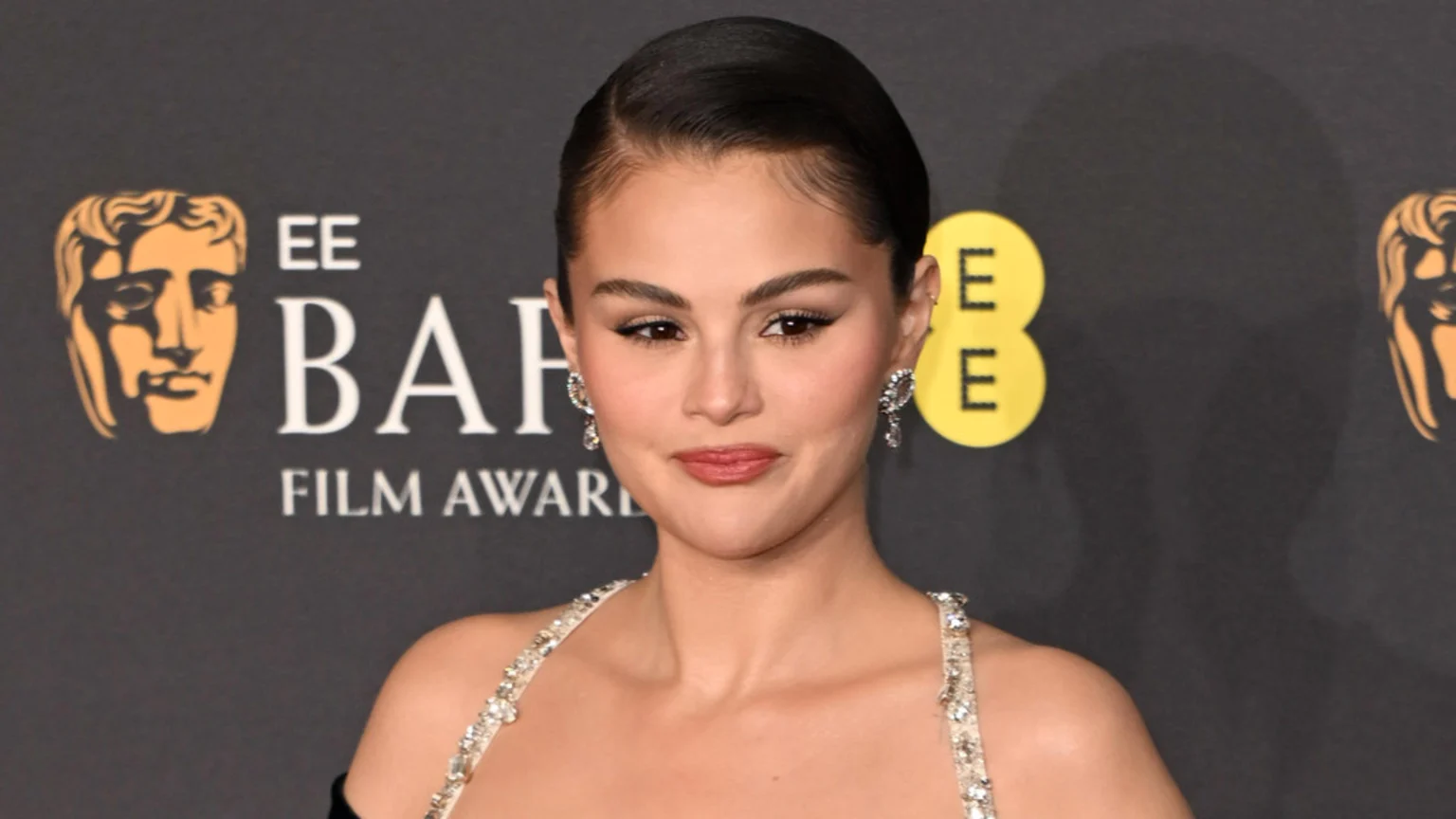 Selena Gomez looks forward with a slight smile, wearing a sleek updo, sparkling earrings, and a light-colored dress with sparkling straps, against a dark backdrop displaying BAFTA EE Film Awards logos.
