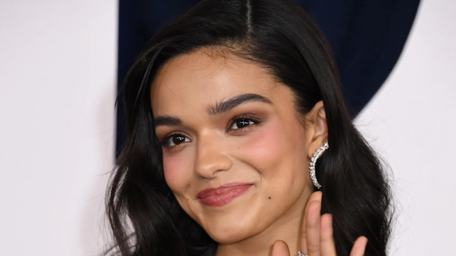 A smiling woman with dark, wavy hair and eyes, wearing a sparkly ear cuff and natural makeup, looks toward the viewer, with a hand and ring visible.