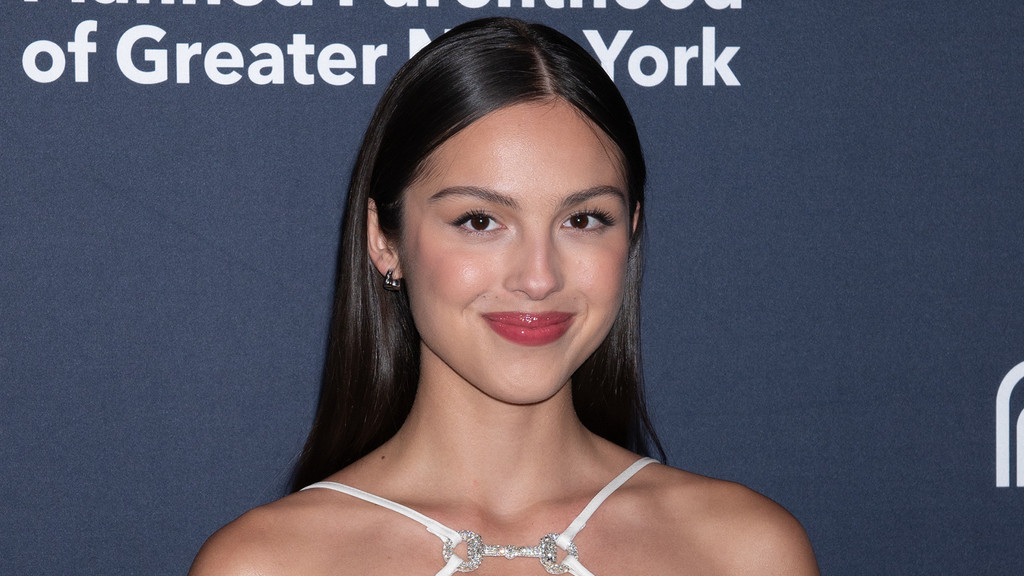 A young woman with long dark hair parted in the middle smiles slightly at the camera, wearing a light-colored top with a sparkling neckline against a dark blue background with partial white text.