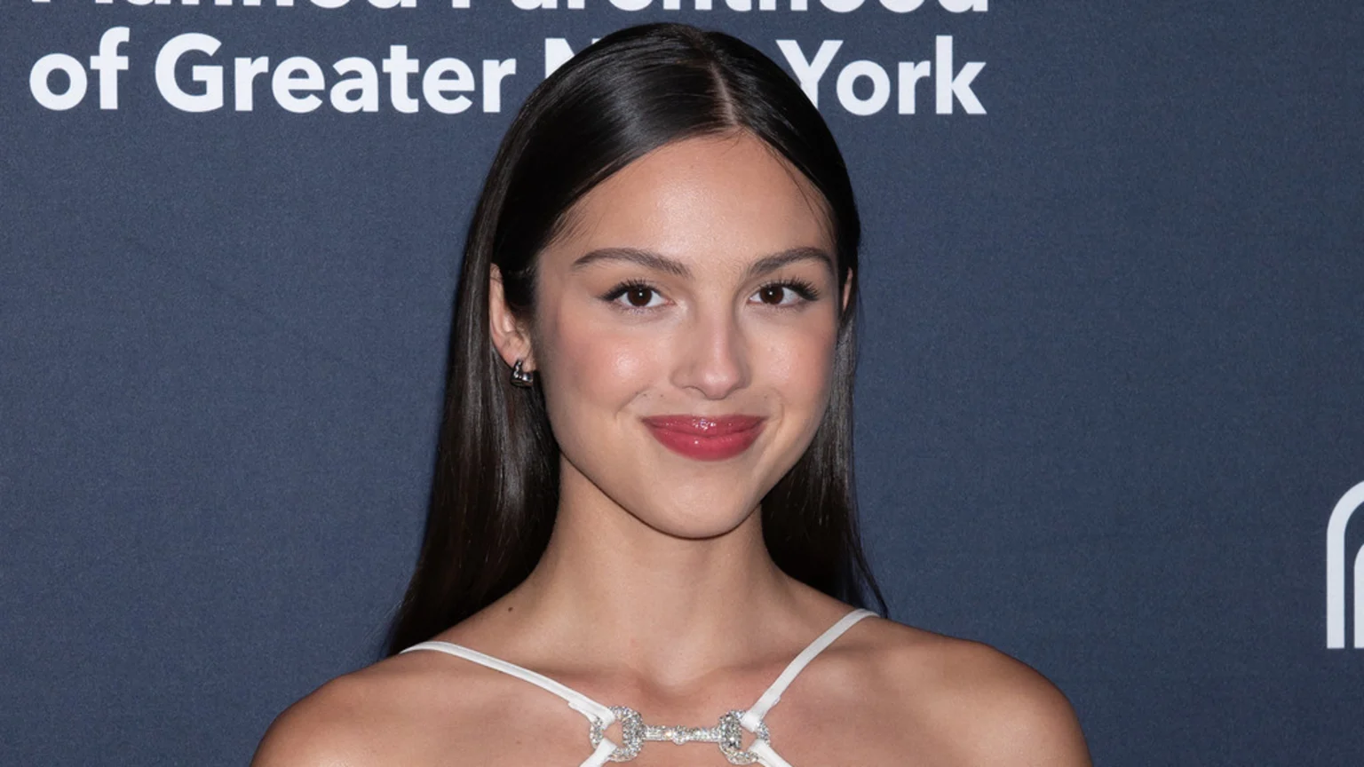 A young woman with long dark hair parted in the middle smiles slightly at the camera, wearing a light-colored top with a sparkling neckline against a dark blue background with partial white text.