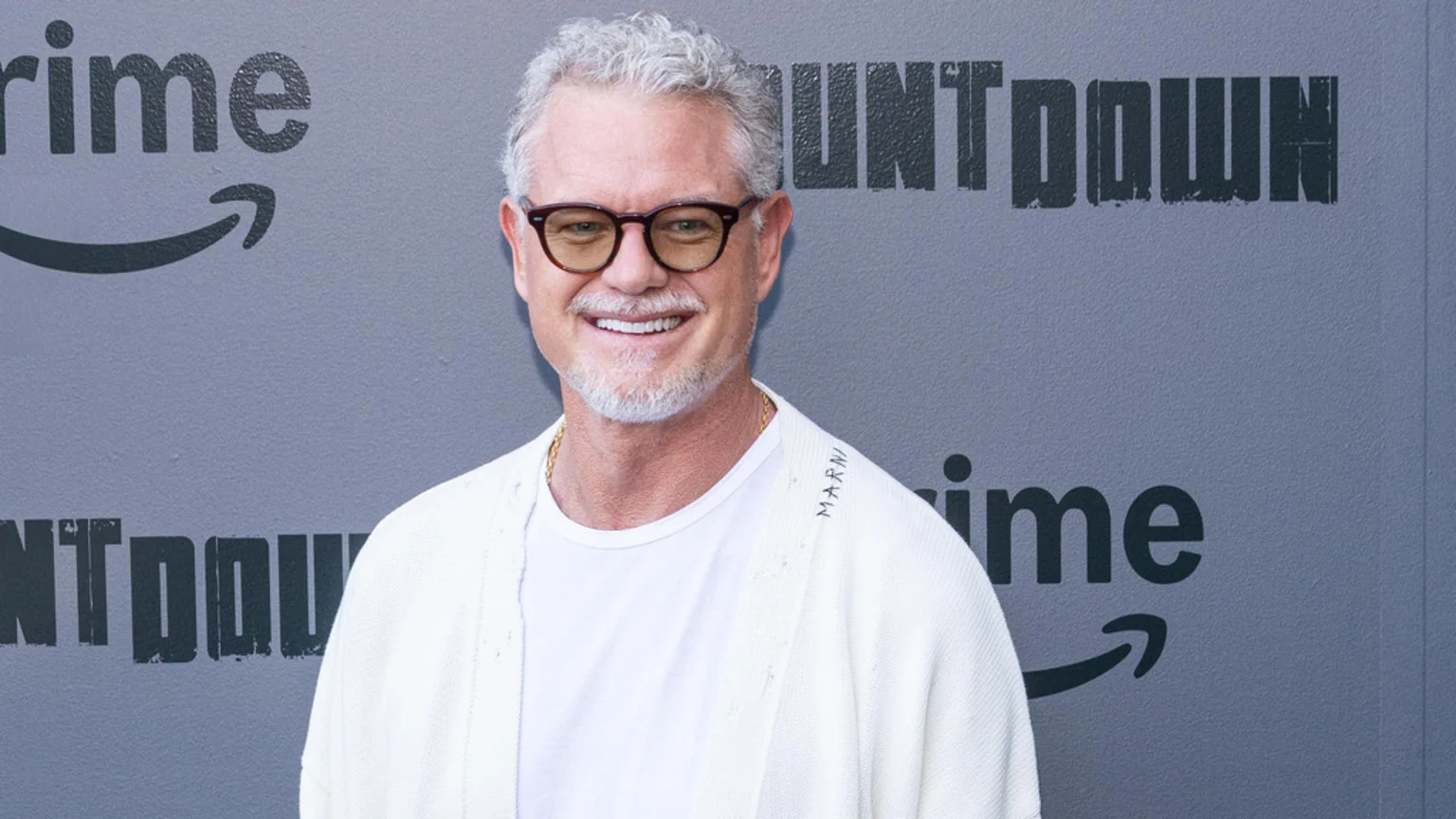 Eric Dane, a smiling man with gray hair and beard, wearing glasses and a white cardigan, stands in front of a gray wall with Amazon Prime and "COUNTDOWN" logos.