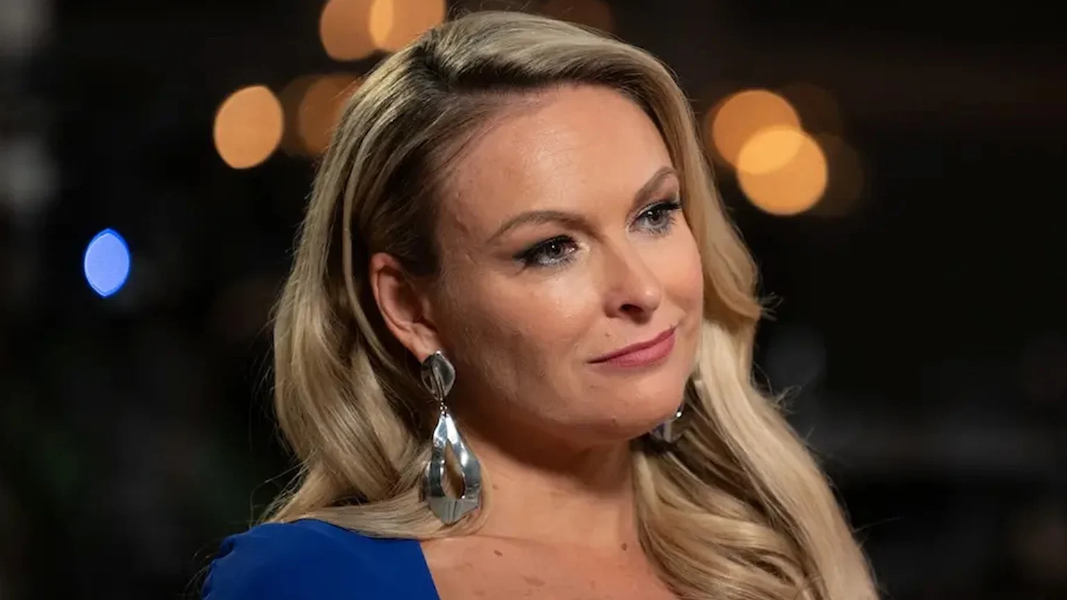 Close-up of Mel Schilling, a blonde woman with blue eyes, wearing silver earrings and a blue top, looking subtly to her right with a slight smile against a blurred dark background.