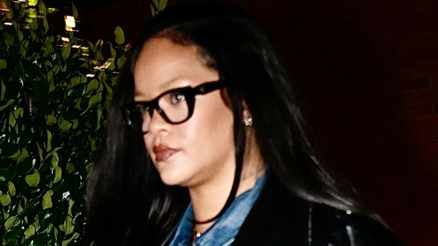 Close-up of Rihanna wearing dark-rimmed glasses, a denim shirt, and a dark jacket, with long dark hair, looking towards the left.