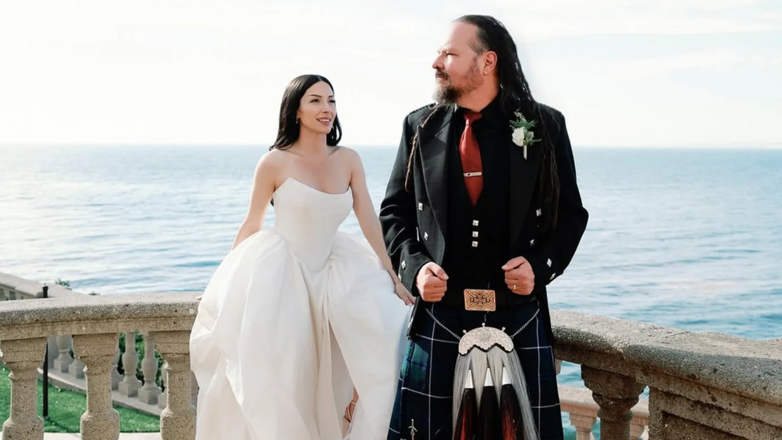 A man with dreadlocks and a beard in a kilt, black jacket, and red tie stands holding hands with a smiling woman in a white strapless wedding gown on a stone balcony overlooking the ocean.
