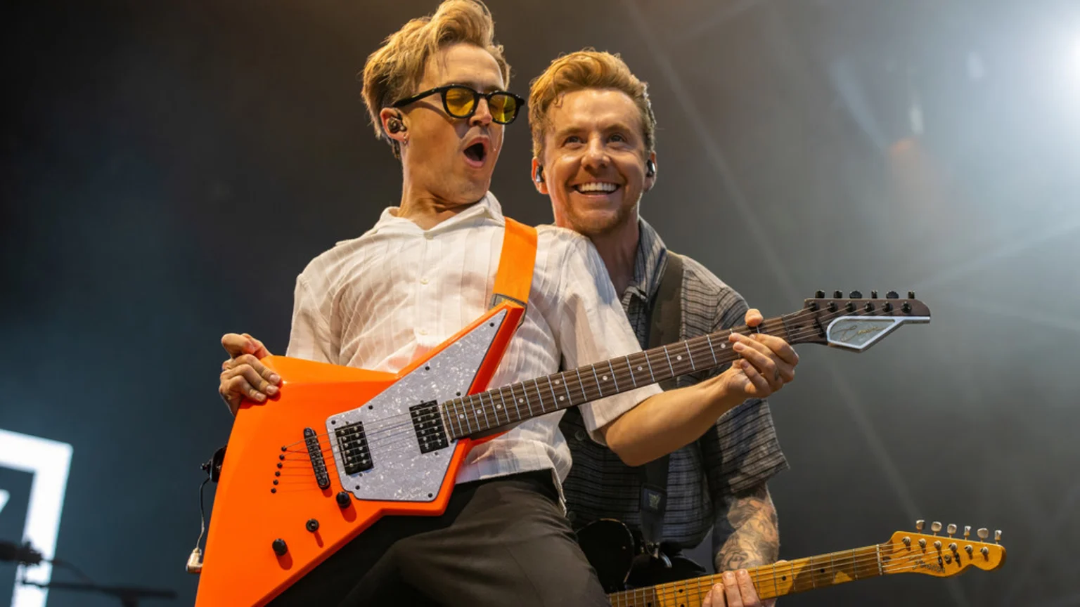 Two male musicians energetically perform on stage; one in yellow sunglasses with an orange V-shaped electric guitar has an open-mouthed expression, while the other behind him smiles while playing his own guitar.