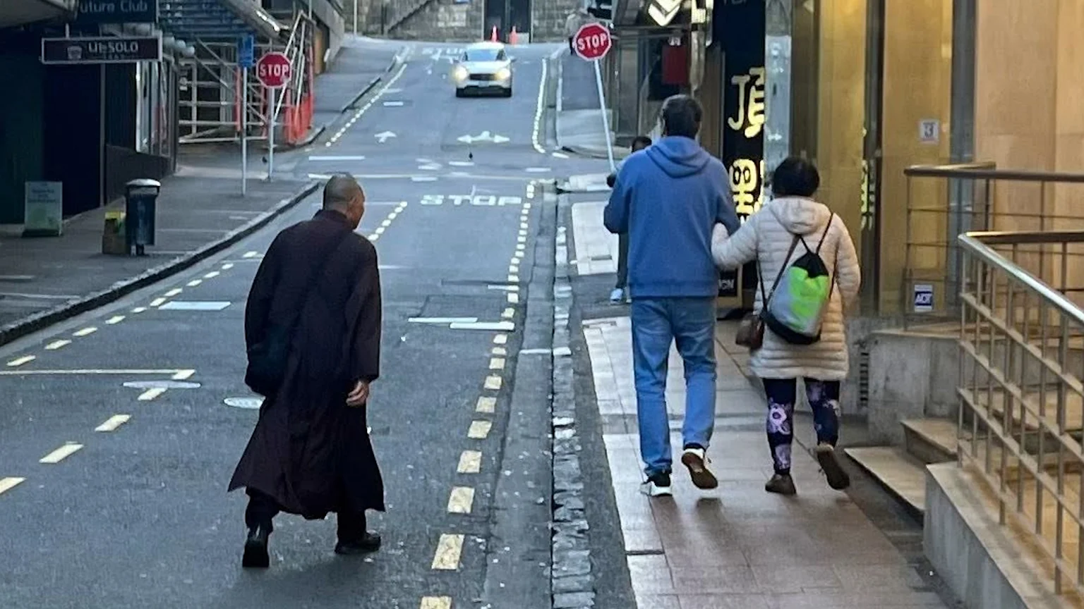 A person in a dark robe walks away on a city street, with a couple holding hands on the sidewalk to their right; a car drives uphill in the background.
