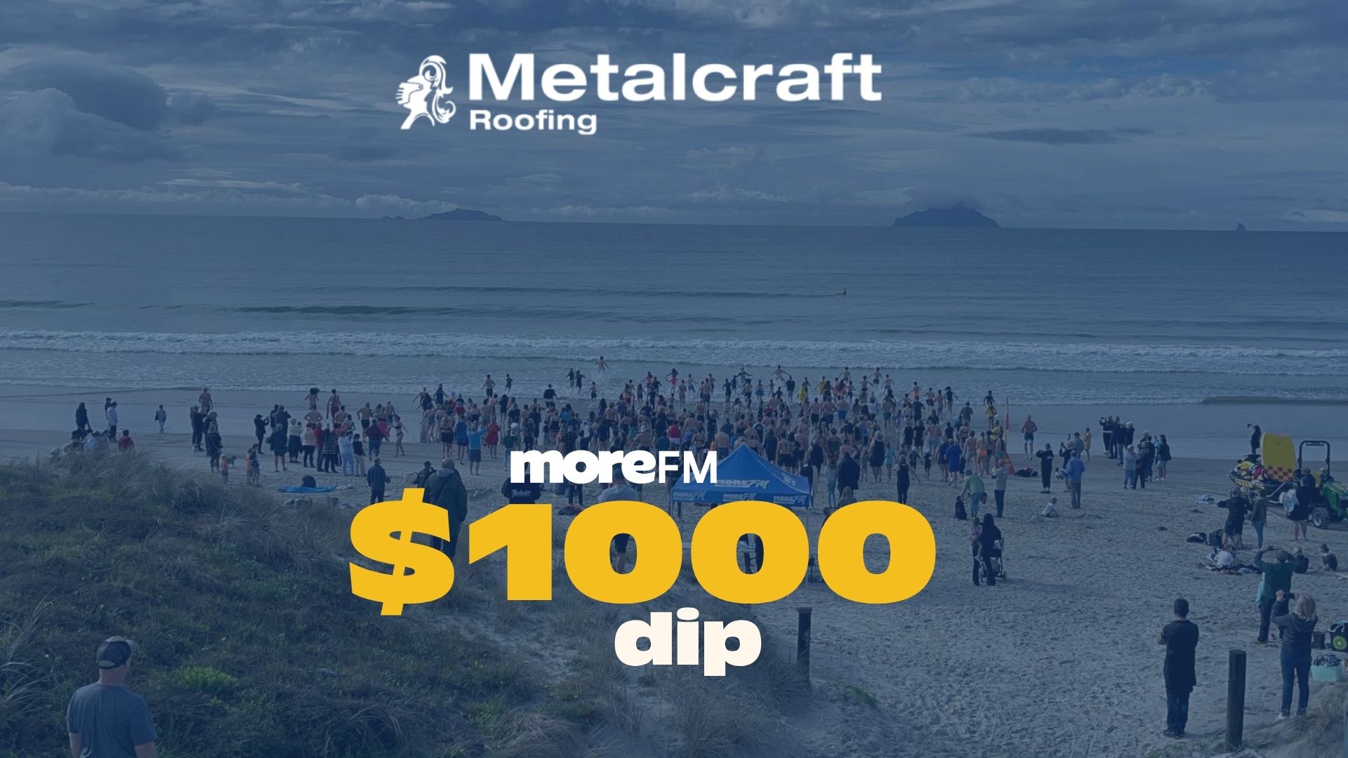 A large crowd of people on a beach are gathered for the moreFM $1000 dip event, with Metalcraft Roofing branding and a lifeguard vehicle visible under a cloudy sky.