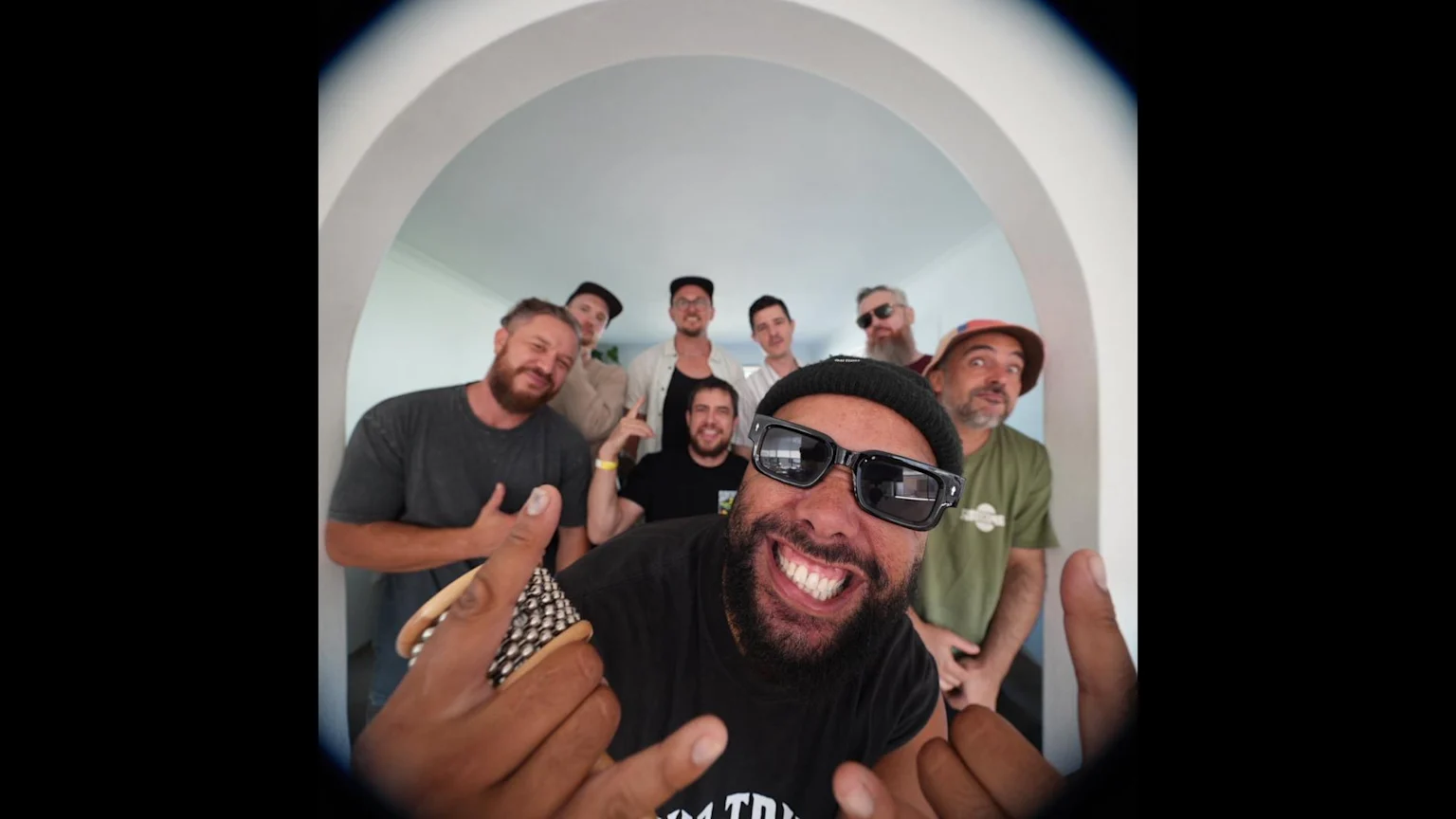 Eight men pose in an indoor archway for a fisheye photo, with the foreground man in a beanie and sunglasses smiling widely and holding a small shaker.