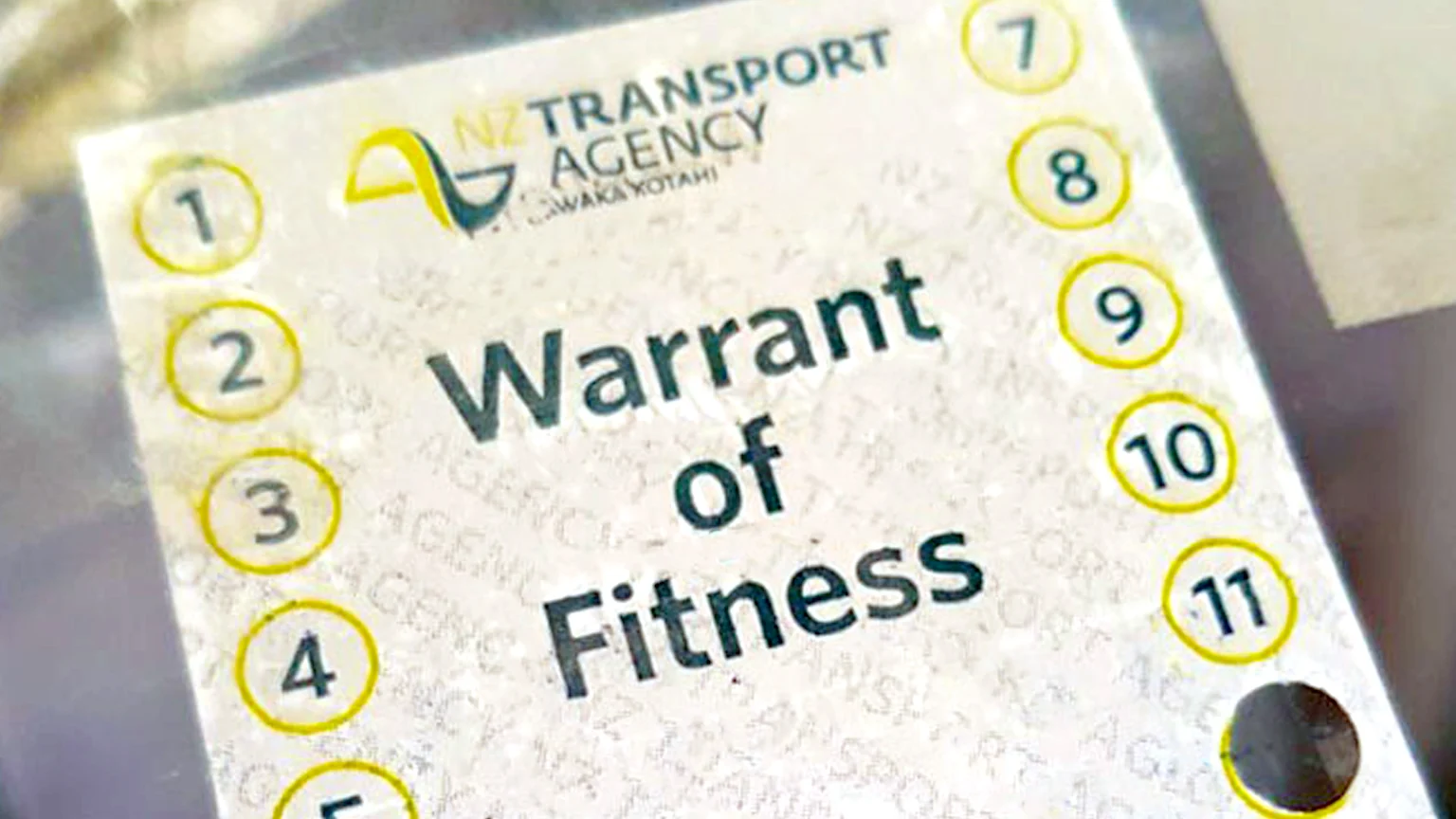 A close-up of a New Zealand Transport Agency "Warrant of Fitness" sticker, showing the agency logo, the title, and numbered circles 1 to 11 with a punched hole.