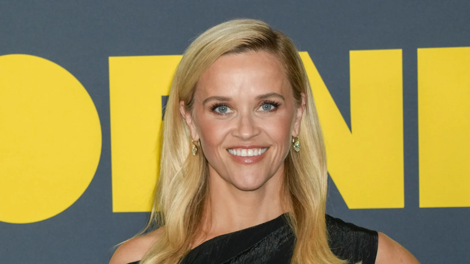 A smiling Reese Witherspoon with long blonde hair, blue eyes, and light makeup wears a black one-shoulder top and earrings in front of a yellow "MORNING" sign.