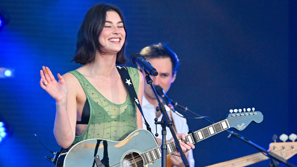 Smiling singer Gracie Abrams, wearing a green top, holds a light blue acoustic guitar and a yellow pick on stage, with a band member visible behind her against a blue background.