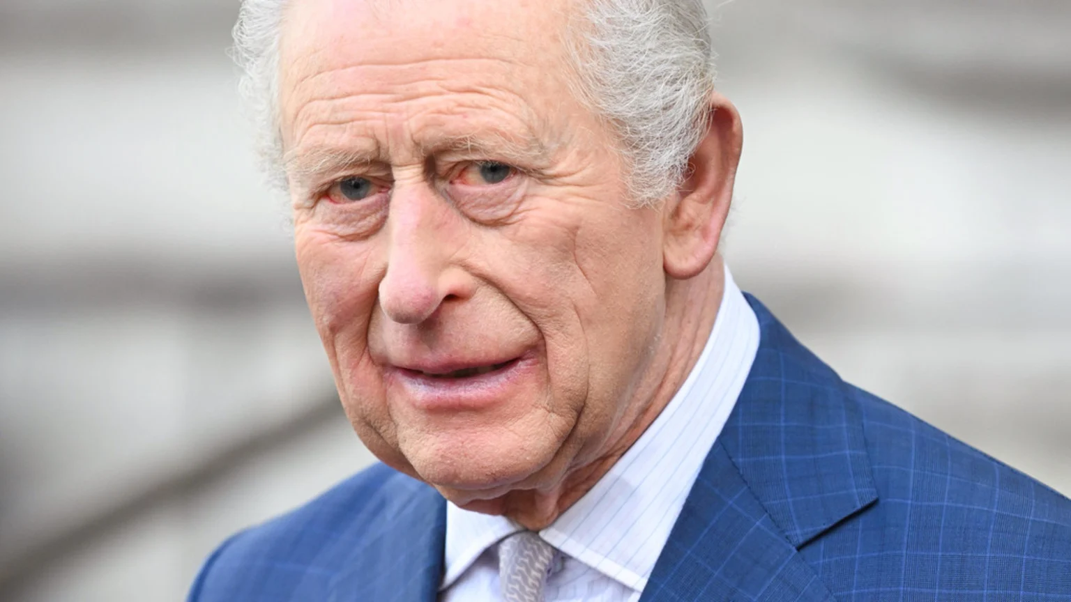 Close-up of King Charles III, an older man with white hair, wearing a blue checkered suit jacket, a light blue striped shirt, and a patterned tie, looking slightly right.