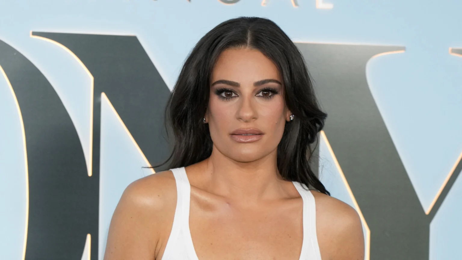 Lea Michele looks directly at the camera, with long dark hair, prominent eye makeup, and a white tank top, against a light blue background with large letters spelling 'TONY'.