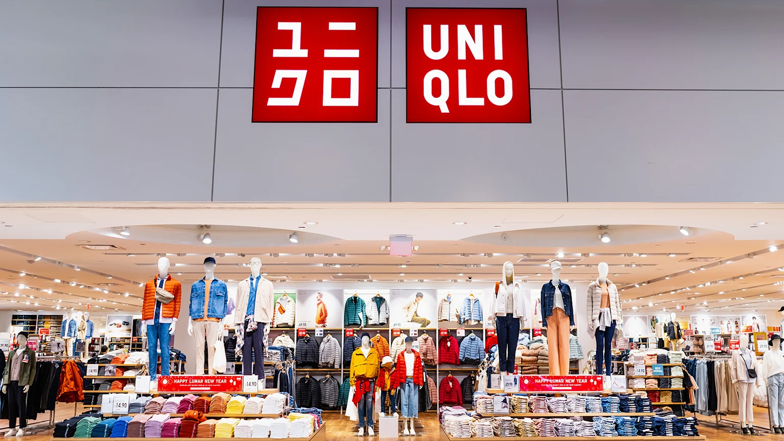 The bright interior of a Uniqlo store featuring large red signs with Japanese characters and the UNIQLO logo, mannequins displaying a range of clothing, and neatly stacked clothes on display, with some "Happy Lunar New Year" banners.