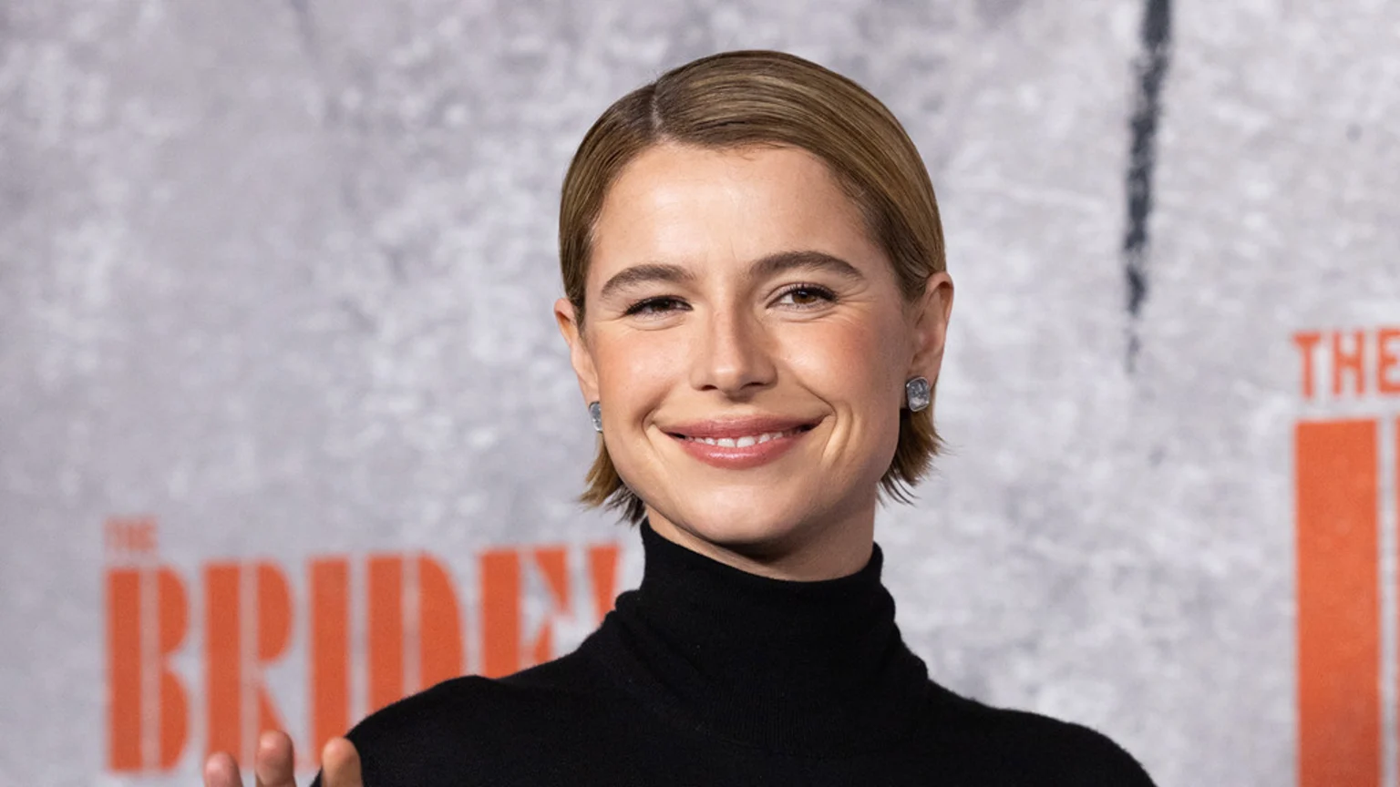 Jessie Buckley smiles directly at the camera, wearing a black turtleneck and stud earrings, with her short brown hair neatly styled, against a blurry gray background with orange text.