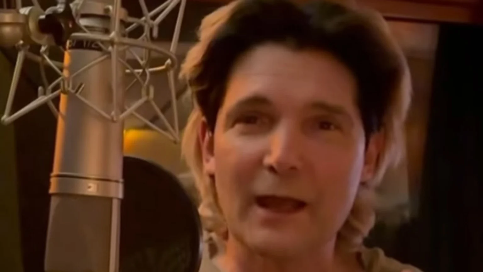 A man with light brown hair looks directly at the camera, mouth slightly open, standing next to a large recording microphone in a shock mount in a warmly lit room.
