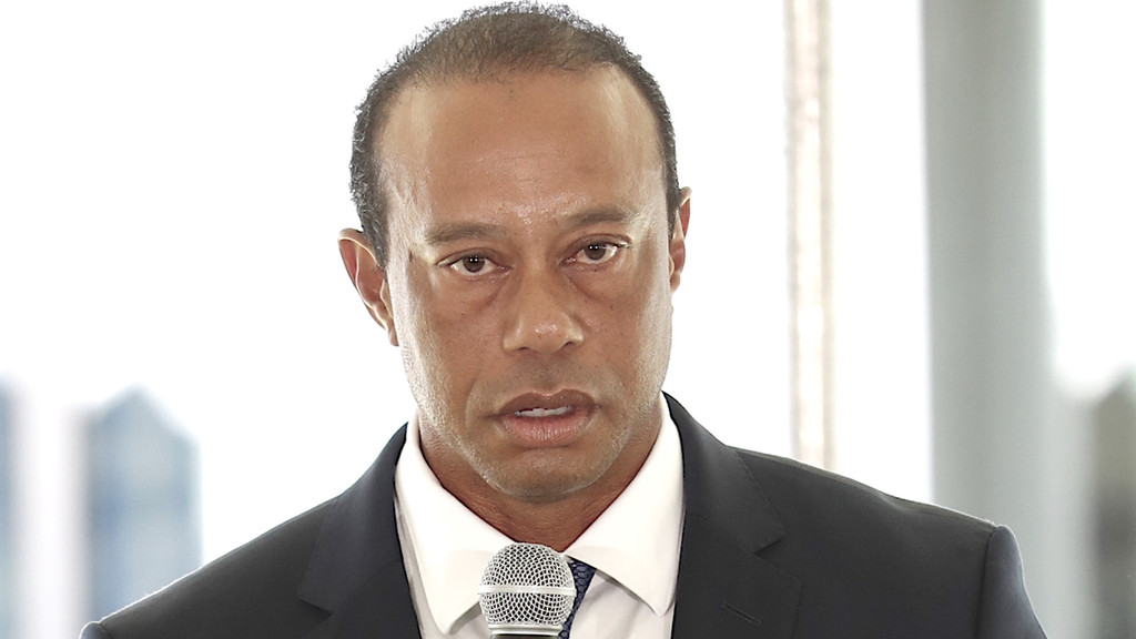 Tiger Woods speaks into a microphone, wearing a dark suit, white shirt, and patterned tie, against a bright, blurred background.
